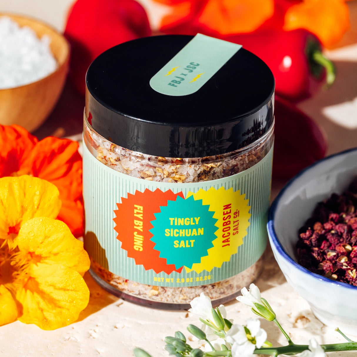Jar of JSC x Fly By Jing Tingly Sichuan Salt with colorful label on a table with flowers and spices.