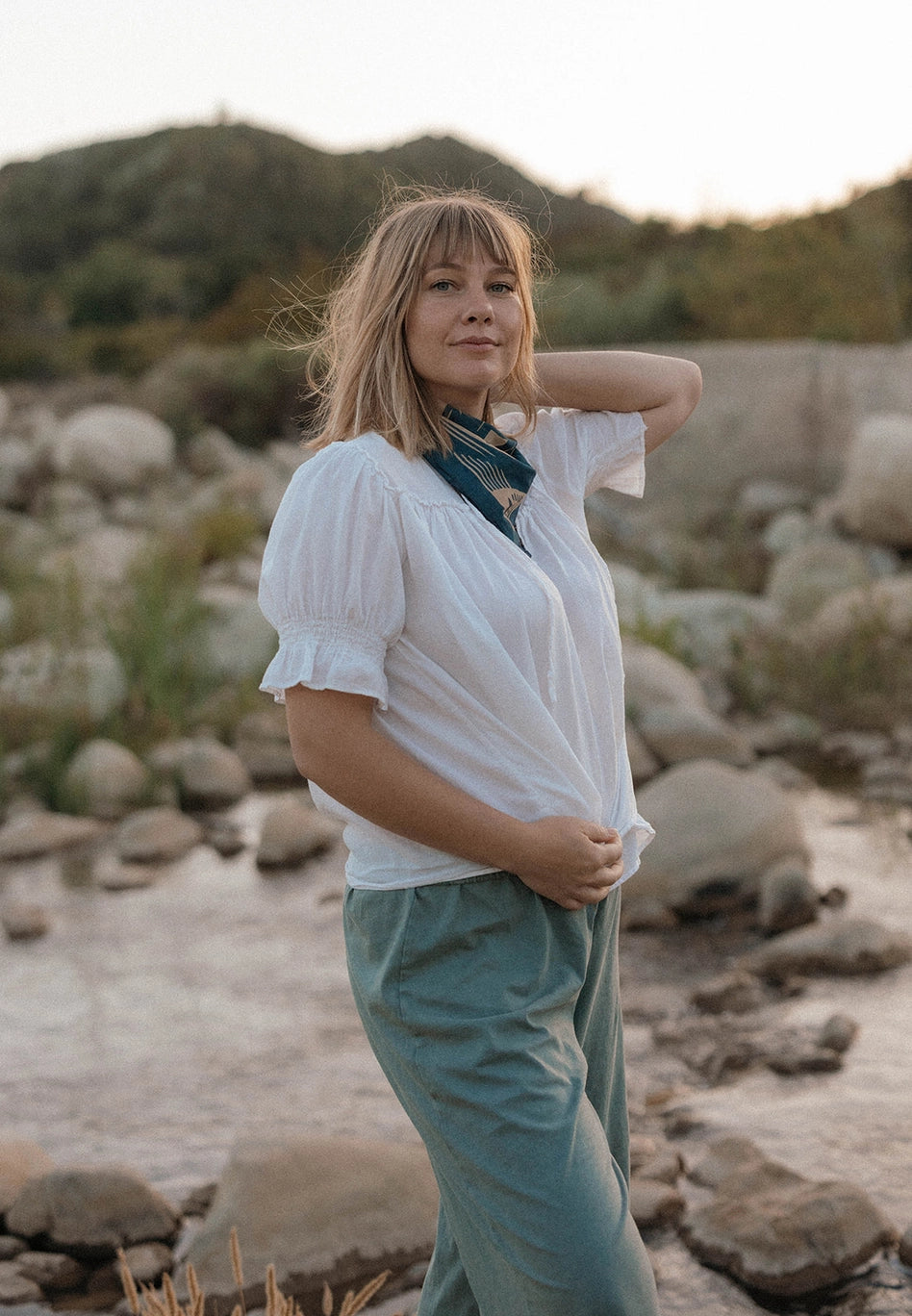 Woman in a white blouse and Bandits El Rito Nights Bandana in a natural setting with rocks and plants.