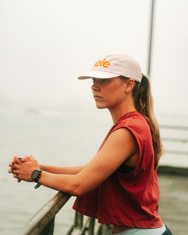 Woman wearing a Ciele Athletics GOCap Comp Century Peachskin pink running cap and a red sleeveless shirt, standing by a body of water.