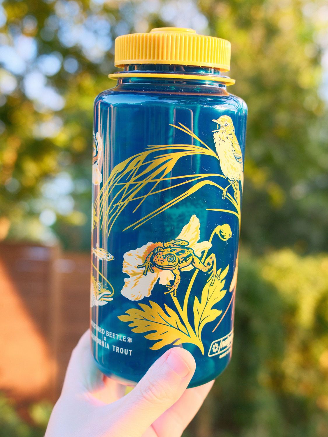 Malibu Creek for CalTrout blue Nalgene water bottle with yellow lid and nature-themed design held in front of a blurred green background