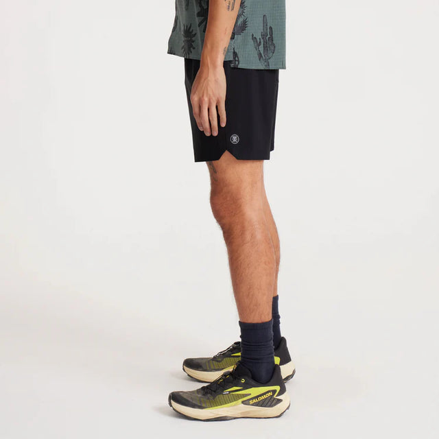 Person wearing Roark Serrano 7" Active Stretch Shorts in Black, navy socks, and green athletic shoes on a white background