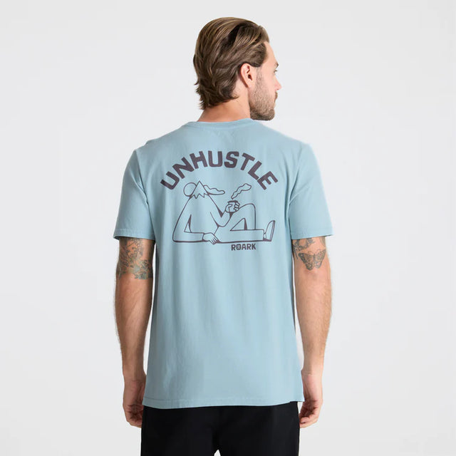 Man wearing a Light blue Roark Unhustle Premium Tee with 'UNHUSTLE' graphic on the back against a white background