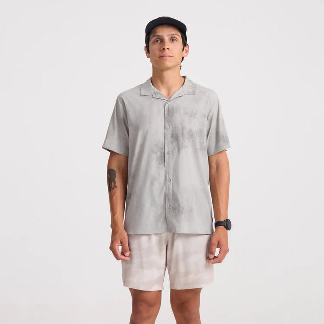 Man wearing a Roark Bless Up Trail Button Up Shirt in Ghost Grey and beige shorts on a white background