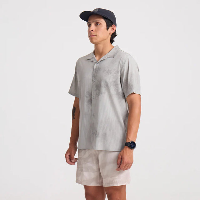 Man wearing a Roark Bless Up Trail Button Up Shirt in Ghost Grey and beige shorts on a white background