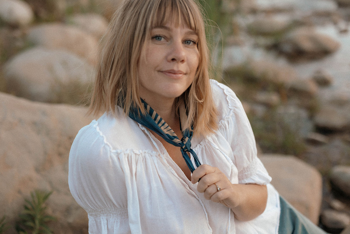 Woman in a white blouse with a Bandits El Rito Nights Bandana standing in a natural setting with rocks and plants.