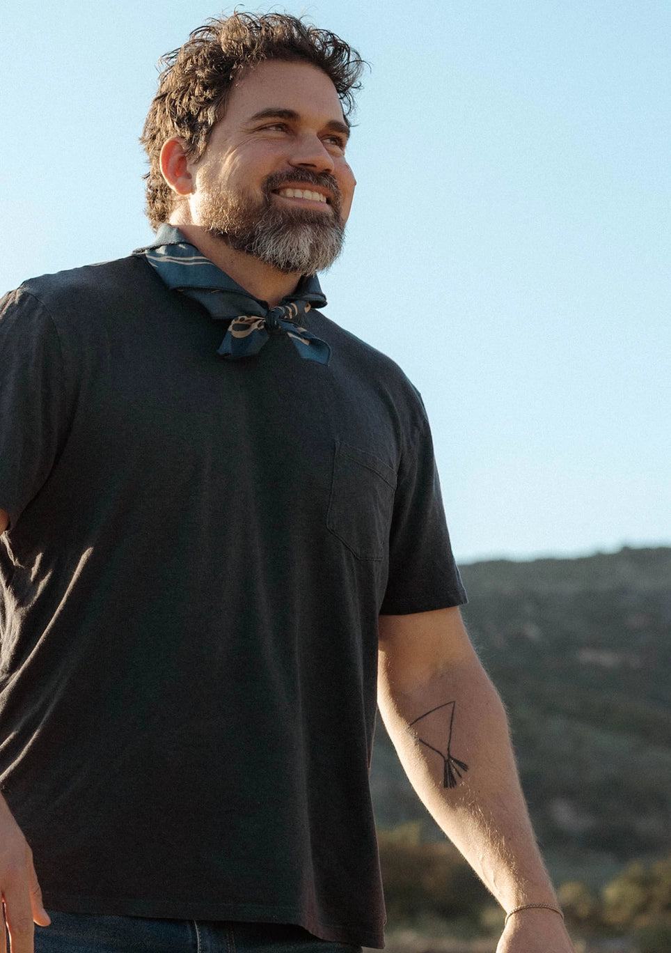 Man wearing a black t-shirt with a Bandits El Rito Nights Bandana around his neck, standing outdoors with a clear sky.