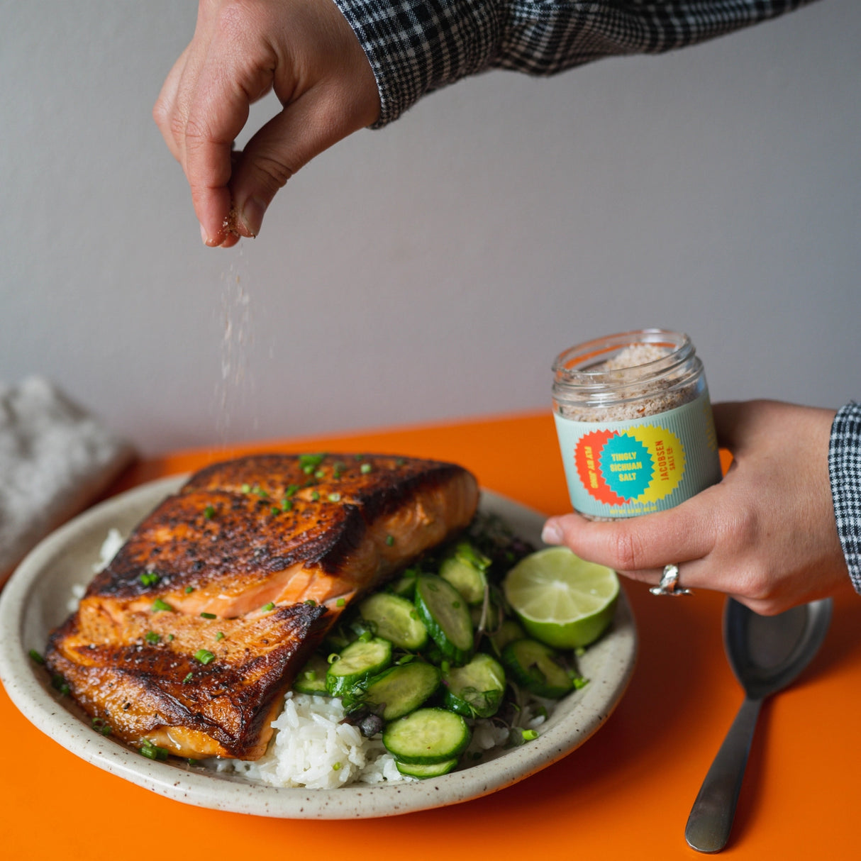 Person seasoning salmon dish with a jar of JSC x Fly By Jing Tingly Sichuan Salt on an orange table.