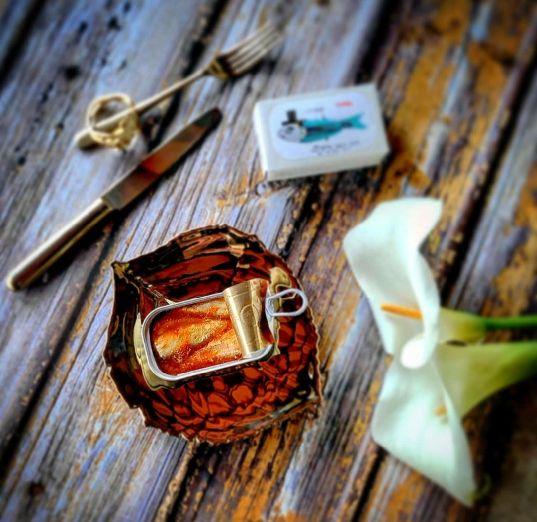 José Gourmet Sardines in Tomato Sauce in a decorative container on a wooden surface with a flower and other items.