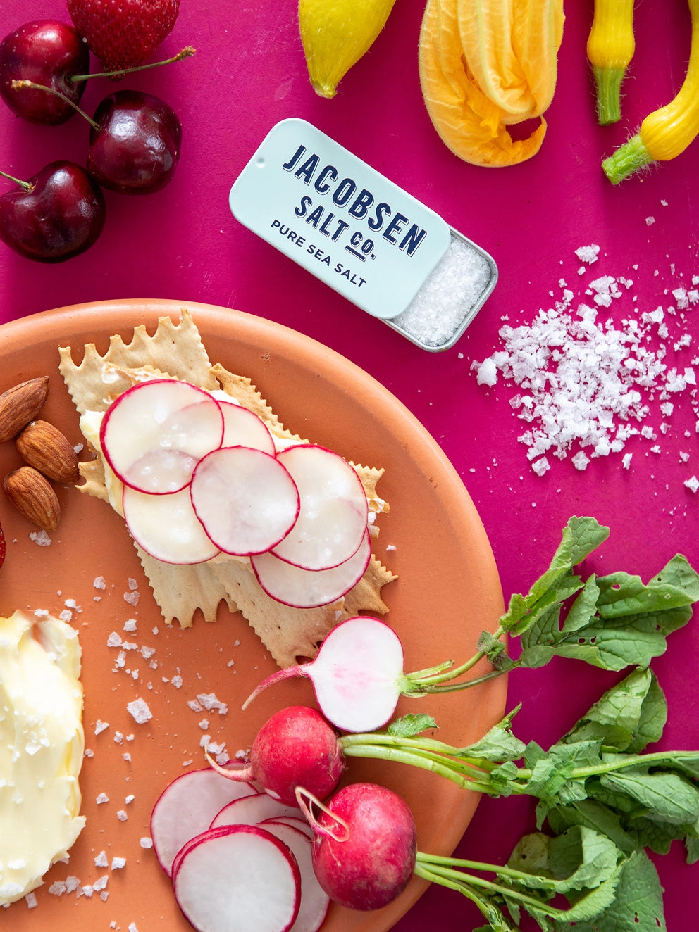 Plate of crackers with radishes, cherry tomatoes, and yellow bell peppers on a pink background with Jacobsen Salt Co. Kosher Sea Salt Slide Tin.