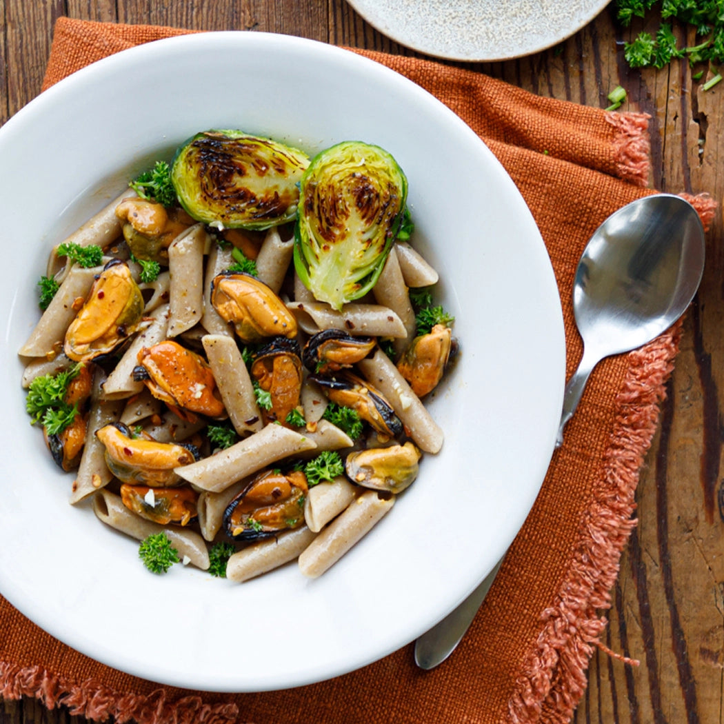 Pasta dish with mussels and brussels sprouts on a white plate with a spoon on an orange napkin.
