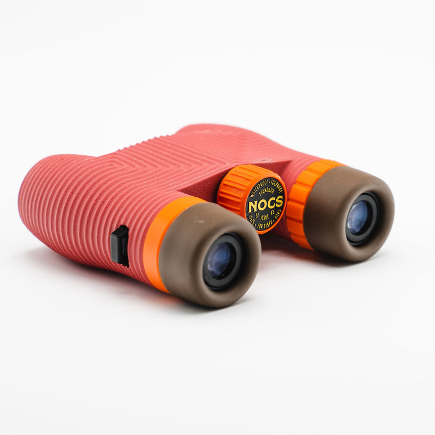 Nocs Standard Issue 10x25 Waterproof Binoculars in manzanita red at an angle on a neutral background.