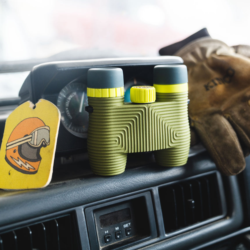 Nocs Standard Issue 10x25 Waterproof Binoculars in olive green on the dashboard of a car.