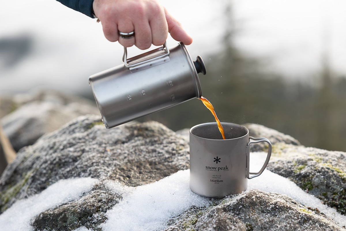 Snow Peak Titanium French Press and Titanium Double Wall Mug on a snowy rock outdoors.