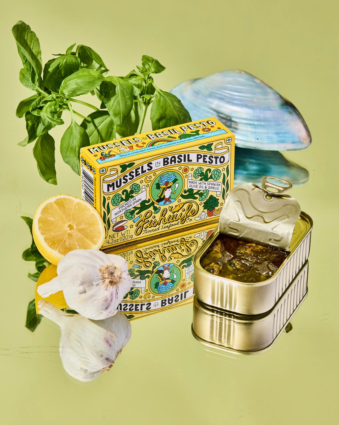 Fishwife Mussels in Basil Pesto can with basil, lemon, and garlic on a green background