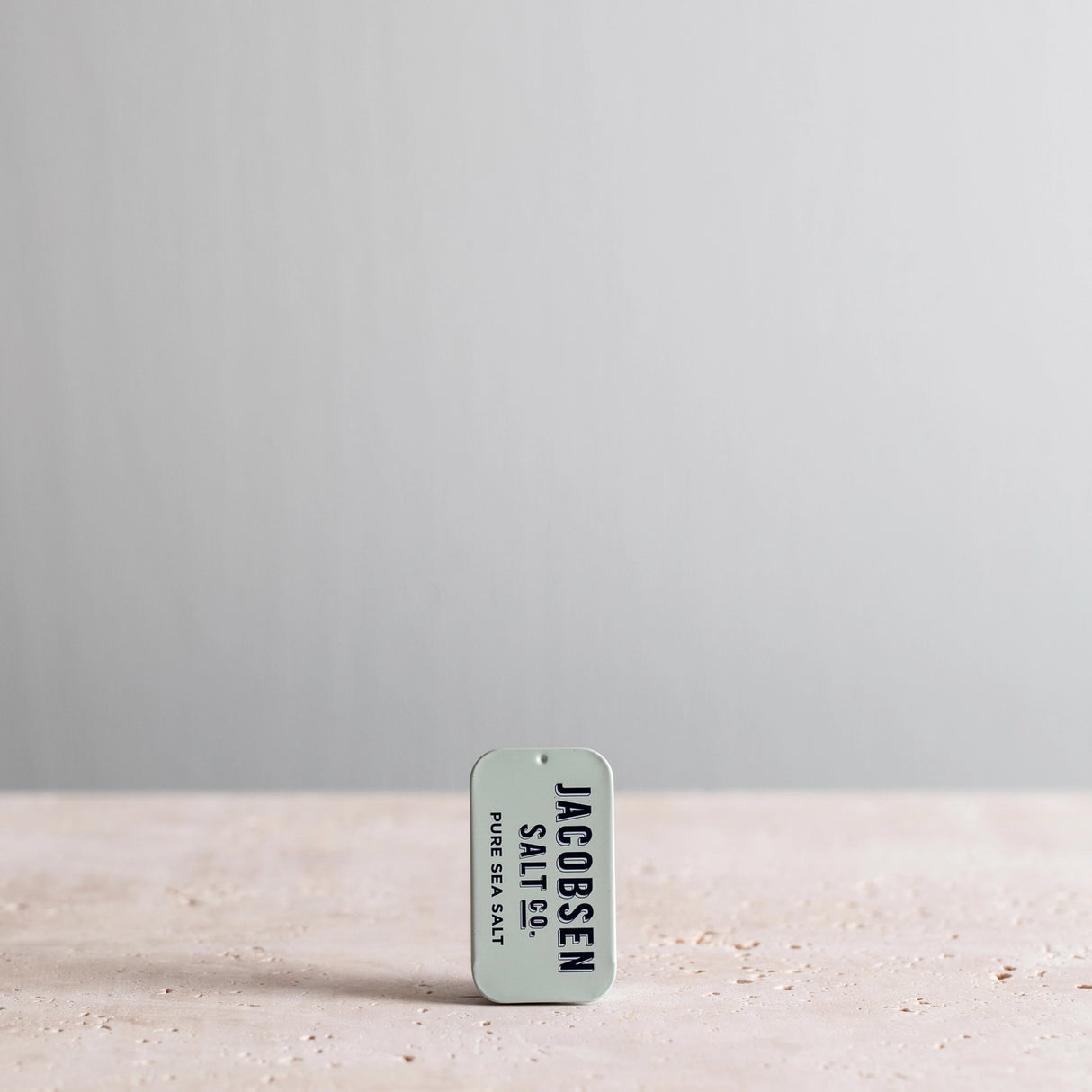 Jacobsen Salt Co. Kosher Sea Salt Slide Tin container on a sandy surface with a gray background