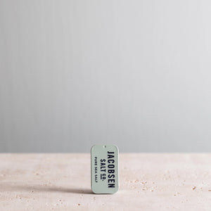 Jacobsen Salt Co. Kosher Sea Salt Slide Tin container on a sandy surface with a gray background