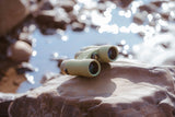 Nocs Provisions Field Issue 10X Waterproof Binoculars in Ponderosa Green outdoors on a rock by the water.