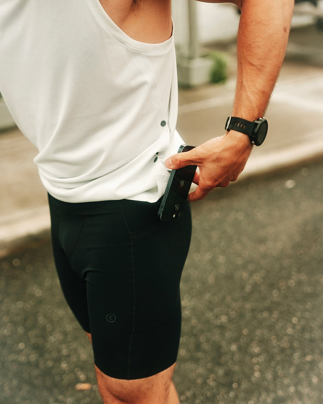 Person wearing a white shirt and Ciele Athletics M FSTTight Short 8" men's running half tight in Shadowcast, holding a phone on a blurred street background