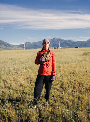 Janji W's Zephyrunner Wind Shell in Nova being worn by a woman from the front outdoors in a field.