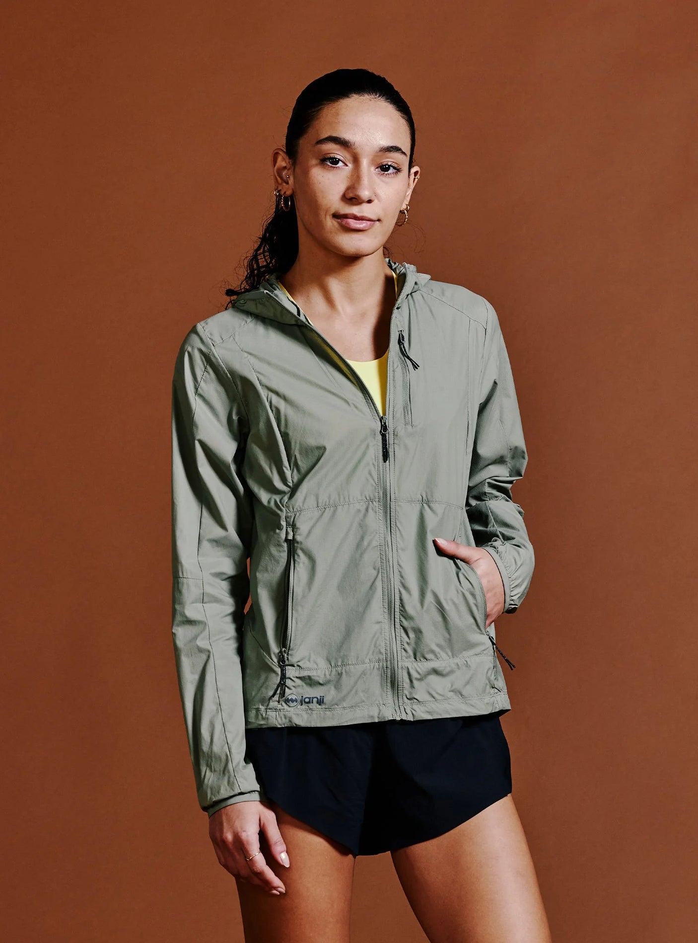 Janji W's Zephyrunner Wind Shell in Sage being worn by a woman from the front on a neutral background.