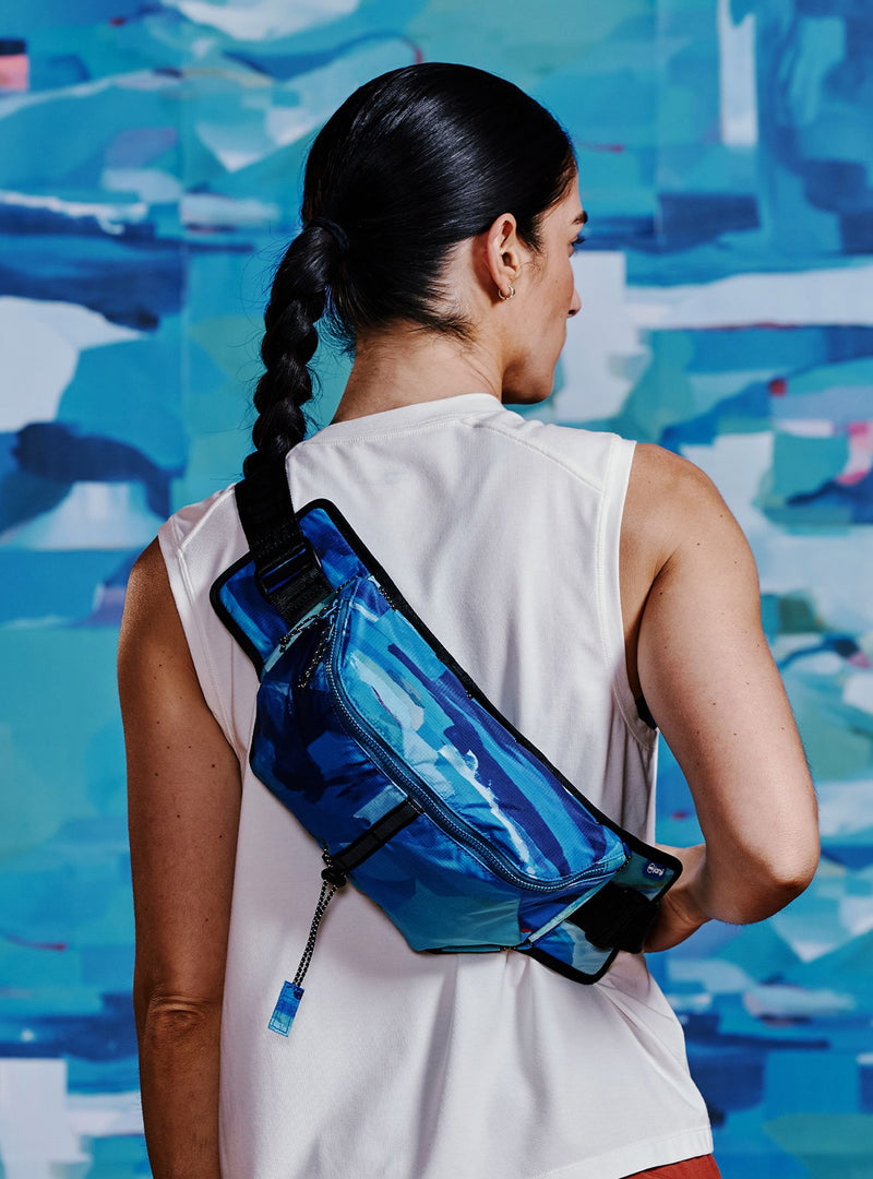 Janji Multipass Sling Bag being worn by a woman against a bold background.