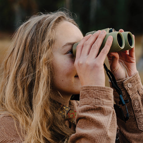 Nocs Provisions Field Issue 10X Waterproof Binoculars in Ponderosa Green being used by a woman outdoors.