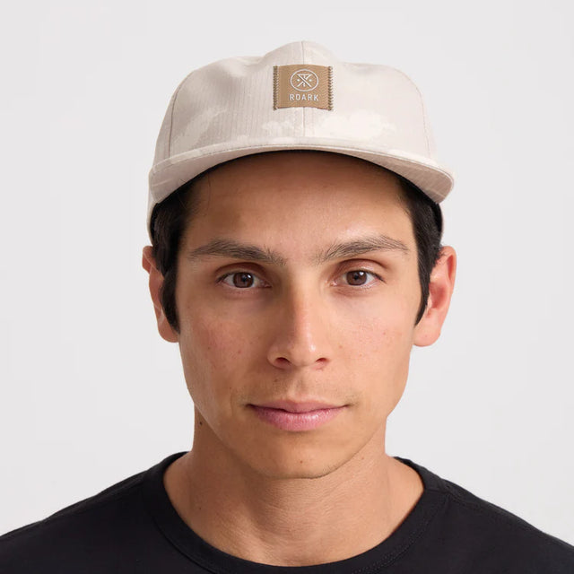 Man wearing a Roark Bless Up Trail Hat Strapback Hat in Fossil with a logo on a light gray background