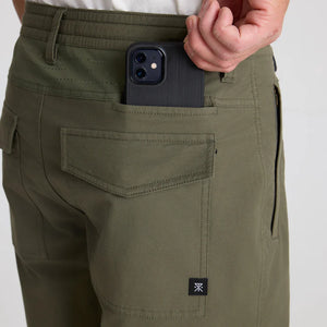 Person wearing Roark Layover Pro Traveler Pants in Military with a phone in the pocket against a neutral background
