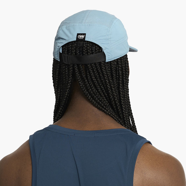 Person wearing a Ciele Athletics GOCap Classic - Athletics Chambray blue running cap with a black strap over their braided hair on a white background