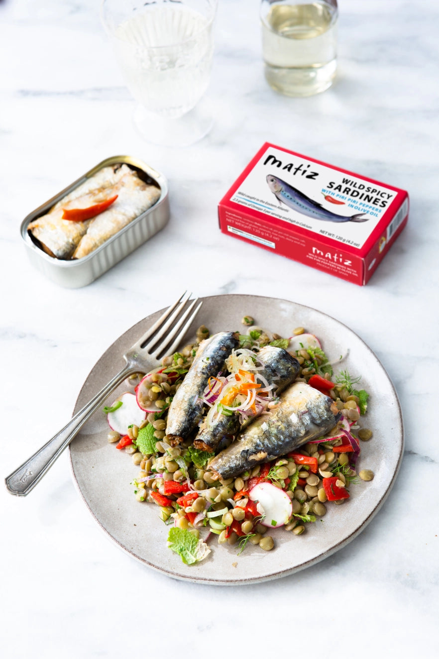 Plate of Matiz Wild Spicy Sardines with Piri Piri Peppers with a side salad next to a box of mafiz sardines on a white surface.