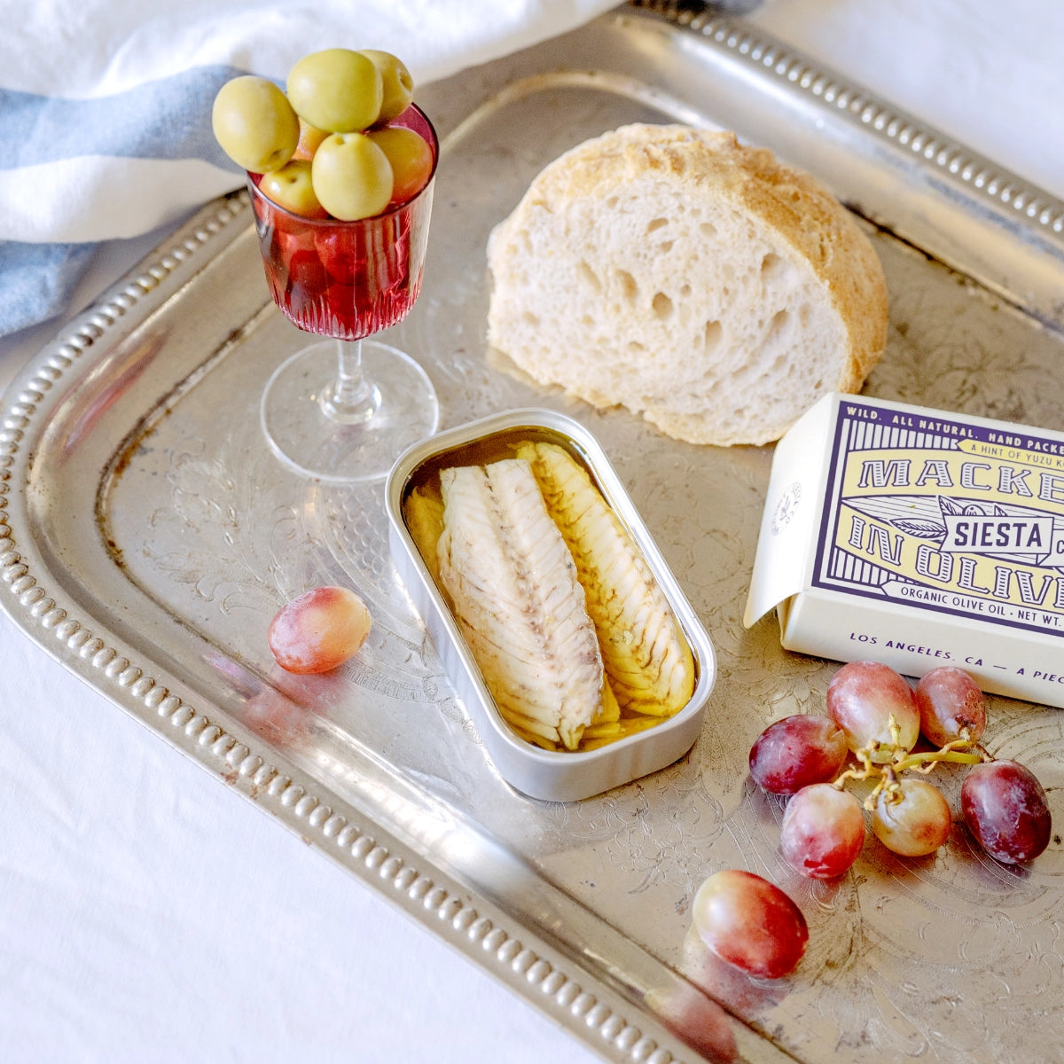 Tray with bread, olives, grapes, and Siesta Co.'s Mackerel in Yuzu Kosho on a white surface.