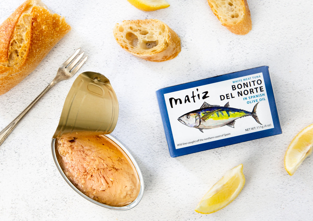 Matiz Bonito del Norte tuna can with open lid, lemon wedges, and bread on a white surface