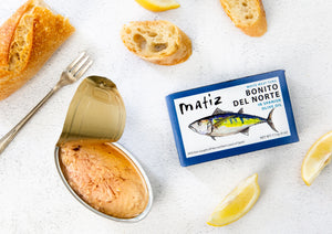 Matiz Bonito del Norte tuna can with open lid, lemon wedges, and bread on a white surface