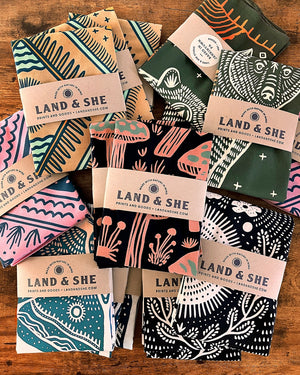 Land & She Folk Song Bandana packaged and shown with other bandanas on a wood background.