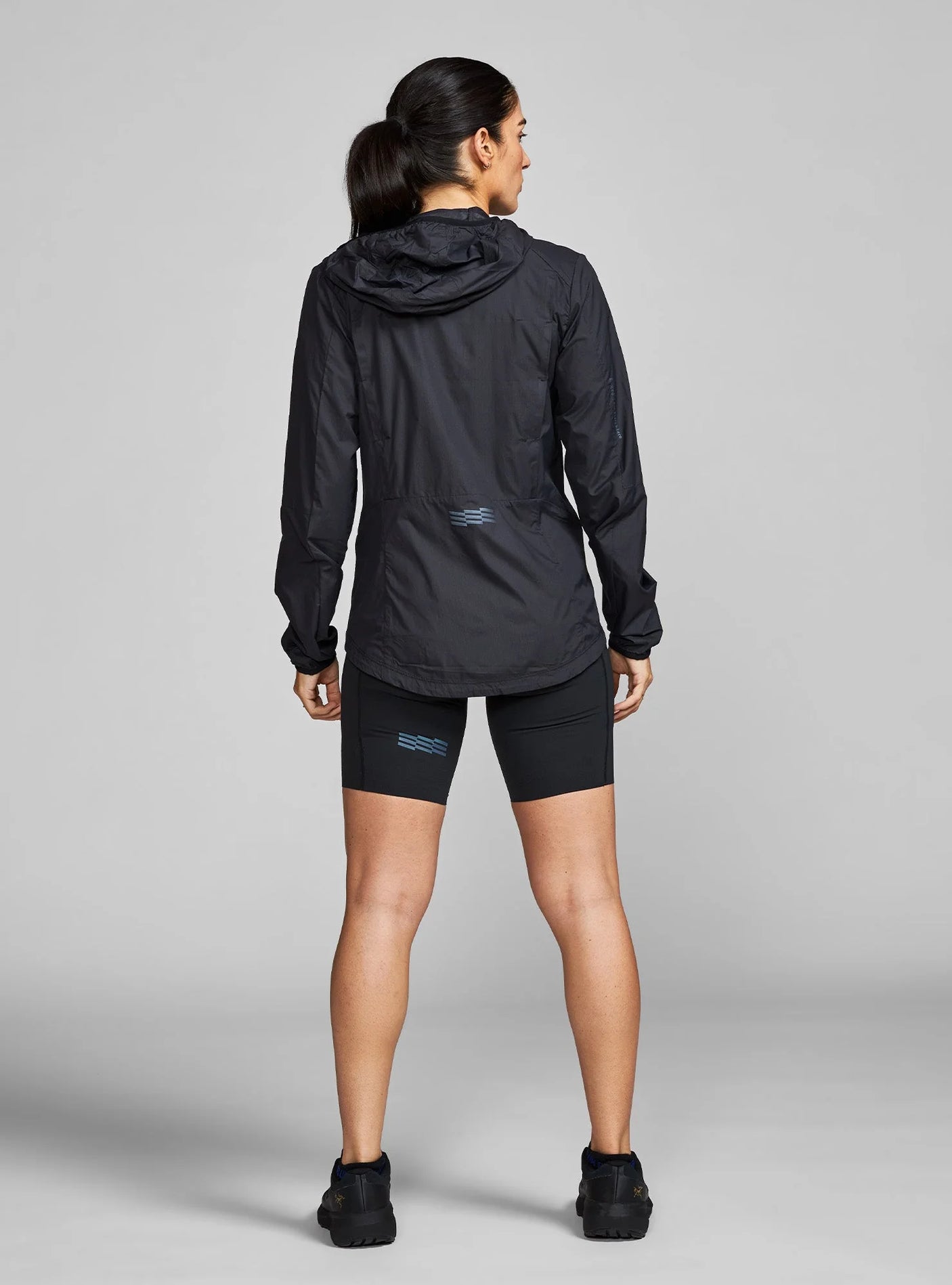 Janji W's Zephyrunner Wind Shell in Midnight being worn by a woman from the rear on a neutral background.