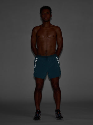 Janji's 5" AFO Middle Short Ultra in Plateau being worn by a man a dark background to show the reflective detailing.