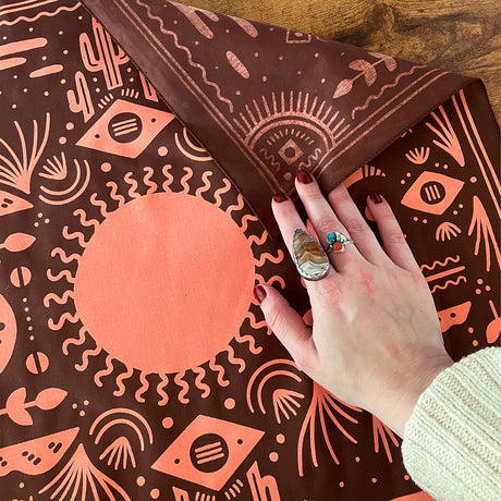 Land & She Western Sun Bandana on a wood background with a woman's hand for comparison.